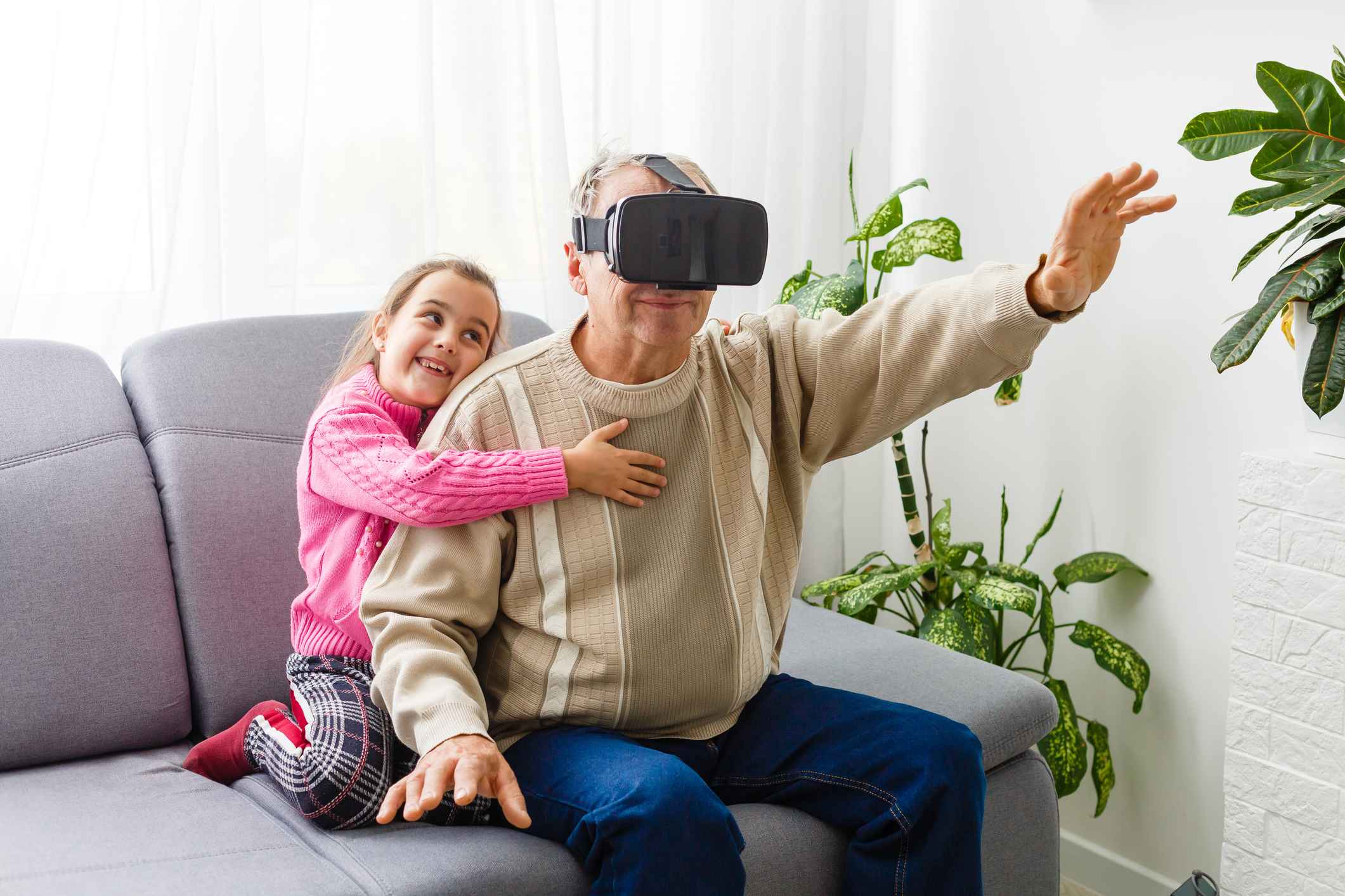 An older man wearing a headset and playing a virutal reality game; his grandchild is seated next to him and embracing him.